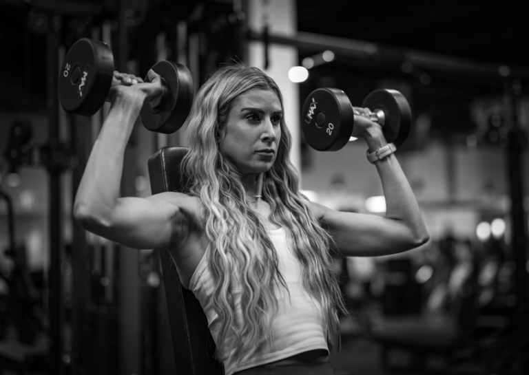 A woman performing shoulder press using dumbbells at the gym, to build a wider shoulder, which gives the illusion of a smaller waist.